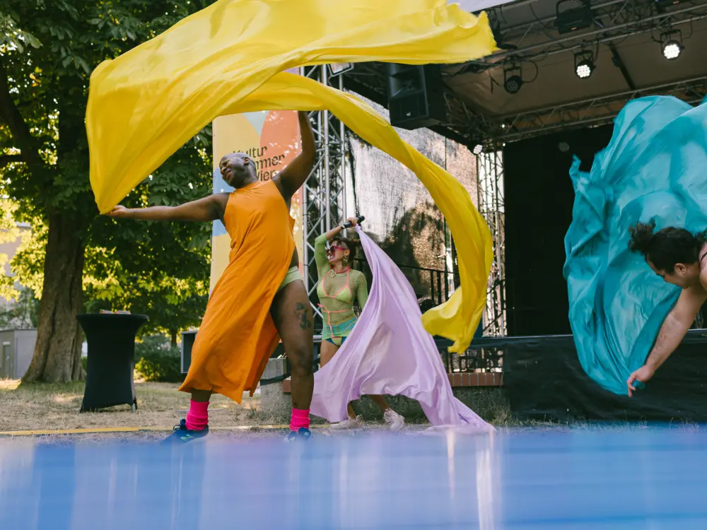 Helena Araújo, Mzamo Nondlwana & Pedra Costa stehen vor der Kultursommer-Bühne im Wilhelmsdorfer Park und schwingen bunte Tücher.