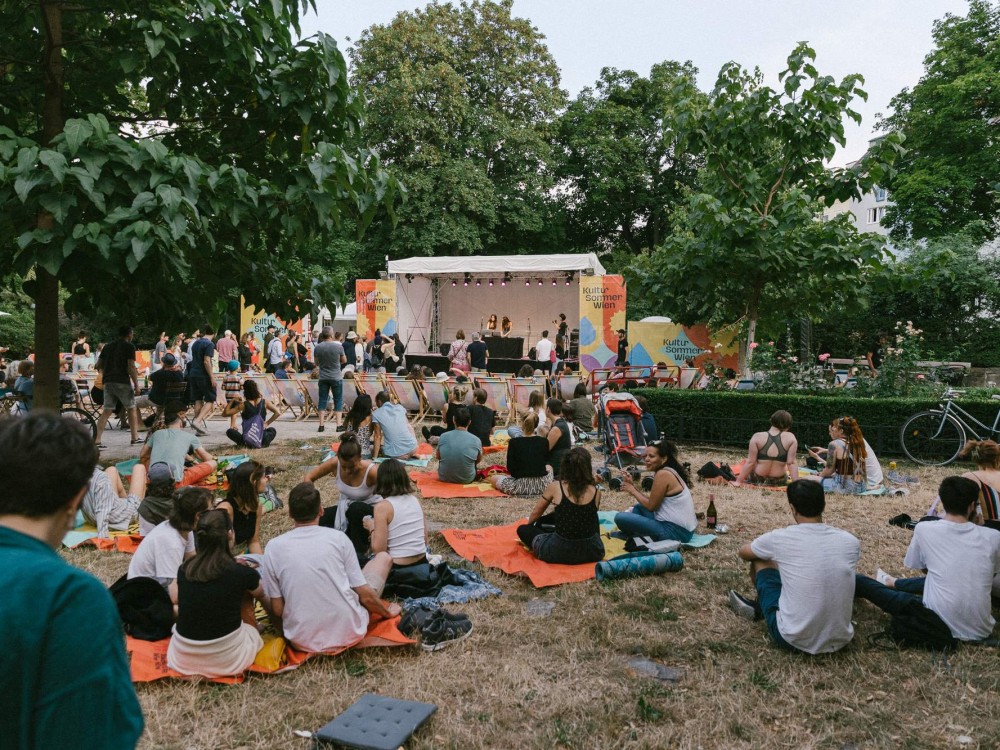 Das Publikum sitzt beim Konzert von Seba Kayan vor der Bühne auf Liegestühlen. Im Vordergrund sitzt das Publikum auf Decken in der Wiese.