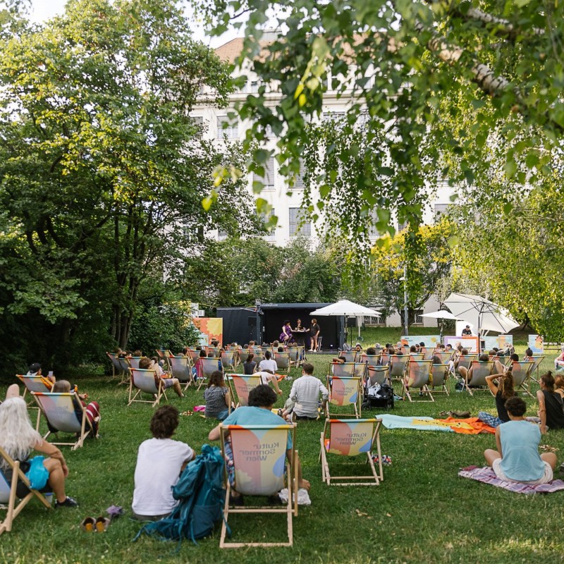 Foto: Theresa Wey Besucherinnen im Wärhingerpark sitzen in Liegestüheln oder auf Picknickdecken in der Wiese und blicken auf die Bühne. 