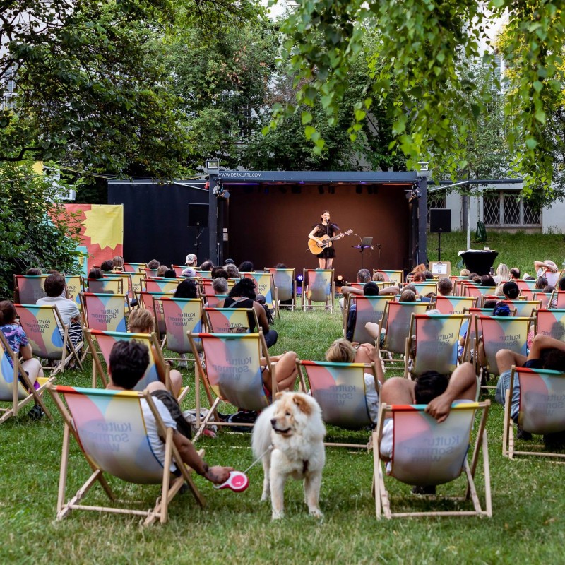 Mira Taylor: Besucherinnen im Wärhingerpark sitzen in Liegestüheln oder auf Picknickdecken in der Wiese und blicken auf die Bühne. Die Musikerin Mira Taylor steht mit Gitarre in der Hand auf der Bühne.