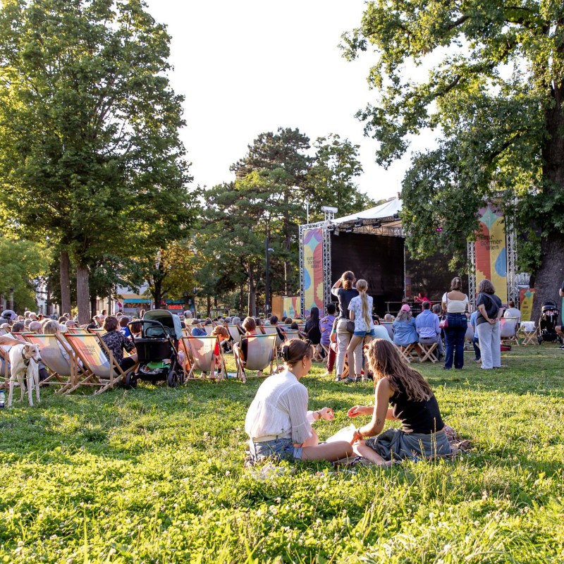 Foto: Judith Stehlik Zwei Personen sitzen etwas abseits der Kultursommer-Bühne in der Wiese. Dahinter sitzen viele Personen in Liegestühlen und schauen dem Programm zu. 