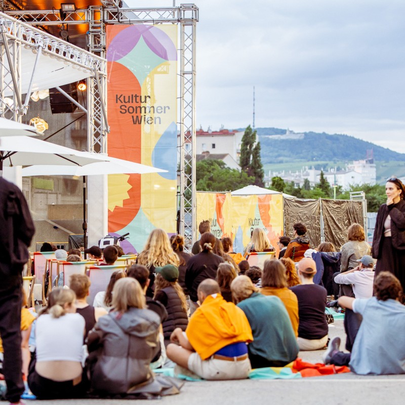 Foto: Judith Stehlik Menschen sitzen und stehen vor der Nordwestbahnhof-Bühne des Kultursommer Wien. Es sind viele Personen im Publikum. 