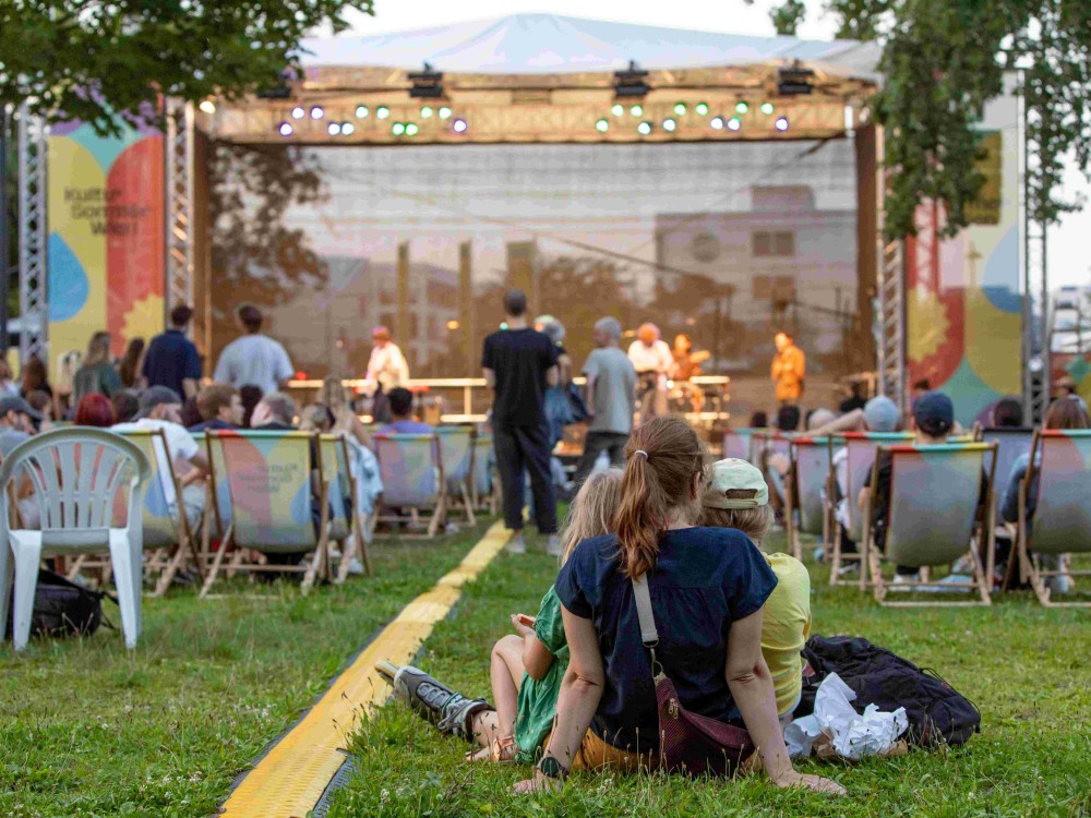 Cordoba78 performt auf der Kultursommer-Bühne im Stadtpark Atzgersdorf. Eine Person sitzt mit zwei Kindern auf der Wiese. 