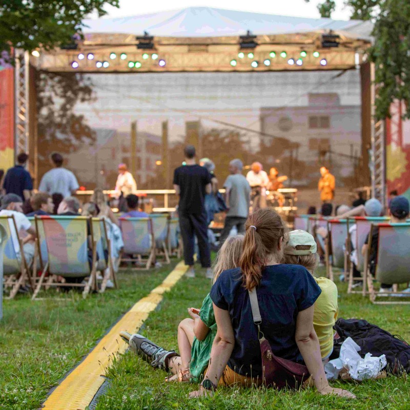 Cordoba78 performt auf der Kultursommer-Bühne im Stadtpark Atzgersdorf. Eine Person sitzt mit zwei Kindern auf der Wiese. 