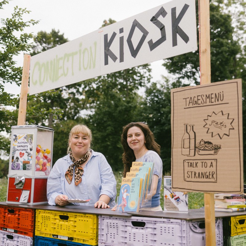 Zwei Personen stehen hinter dem Connection Kiosk-Stand. Auf einem Schild ist eine Illustration vom Tagesmenü. Außerdem steht ein Aufmerksamkeits-Automat auf der Theke. 