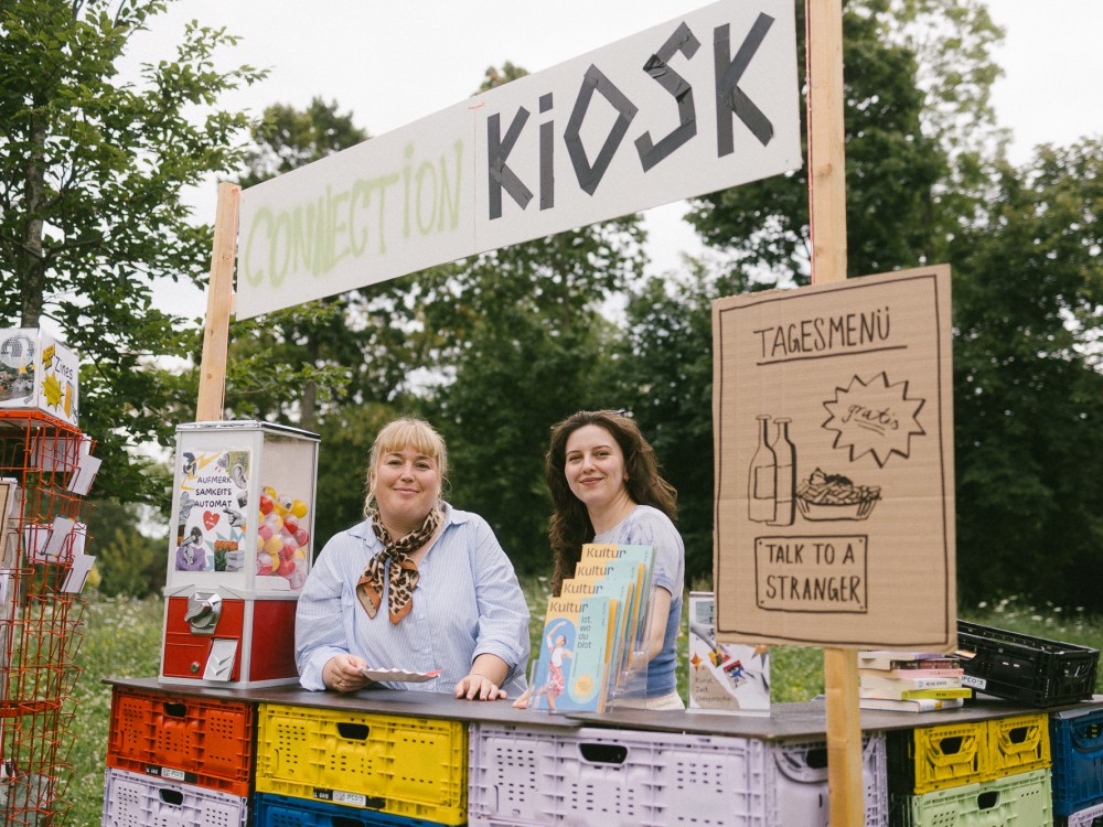 Zwei Personen stehen hinter dem Connection Kiosk-Stand. Auf einem Schild ist eine Illustration vom Tagesmenü. Außerdem steht ein Aufmerksamkeits-Automat auf der Theke. 