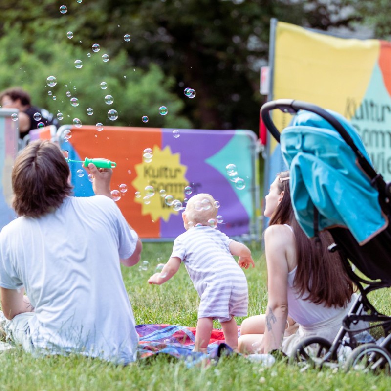 Foto: Theresa Wey Ein Mann und ein Baby sitzen in der Wiese im Währingerpark neben der Bühne des Kultursommers. Der Mann bläst Seifenblasen in die Luft. Daneben steht ein Kinderwagen.