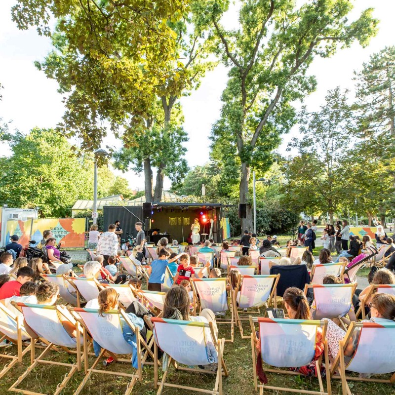 Foto: Judith Stehlik Auftritt von Meo auf der Bühne im Hyblerpark. Man sieht das Publikum von hinten in Liegestühlen sitzend.