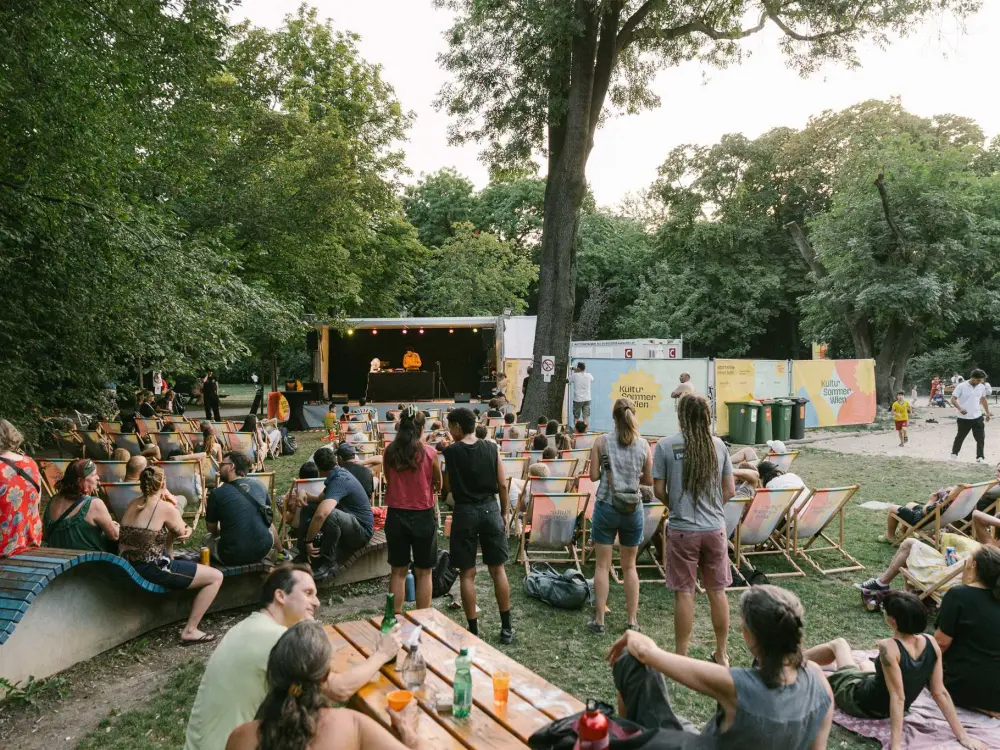 Kimyan performt auf der Kultursommer-Bühne im Waldmüllerpark. Der Park ist gut gefüllt, viele Menschen hören dem Konzert zu.