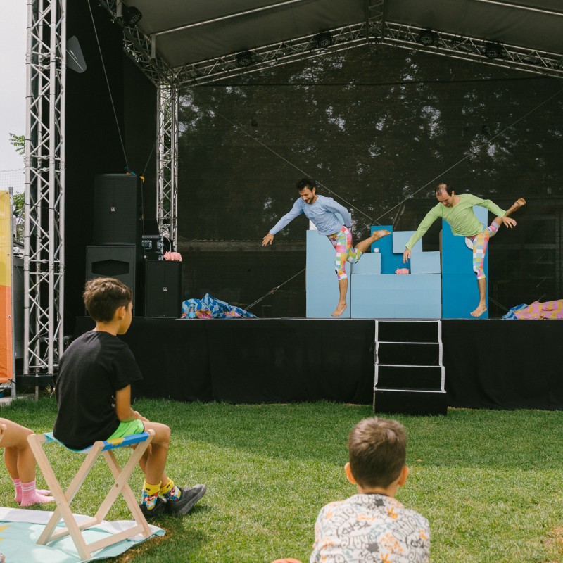 Foto: Niko Havranek BewegKunst performen auf der Kultursommer-Bühne beim Fußballverein 1210. Direkt vor der Bühne sitzen einige Kinder und sehen zu. 