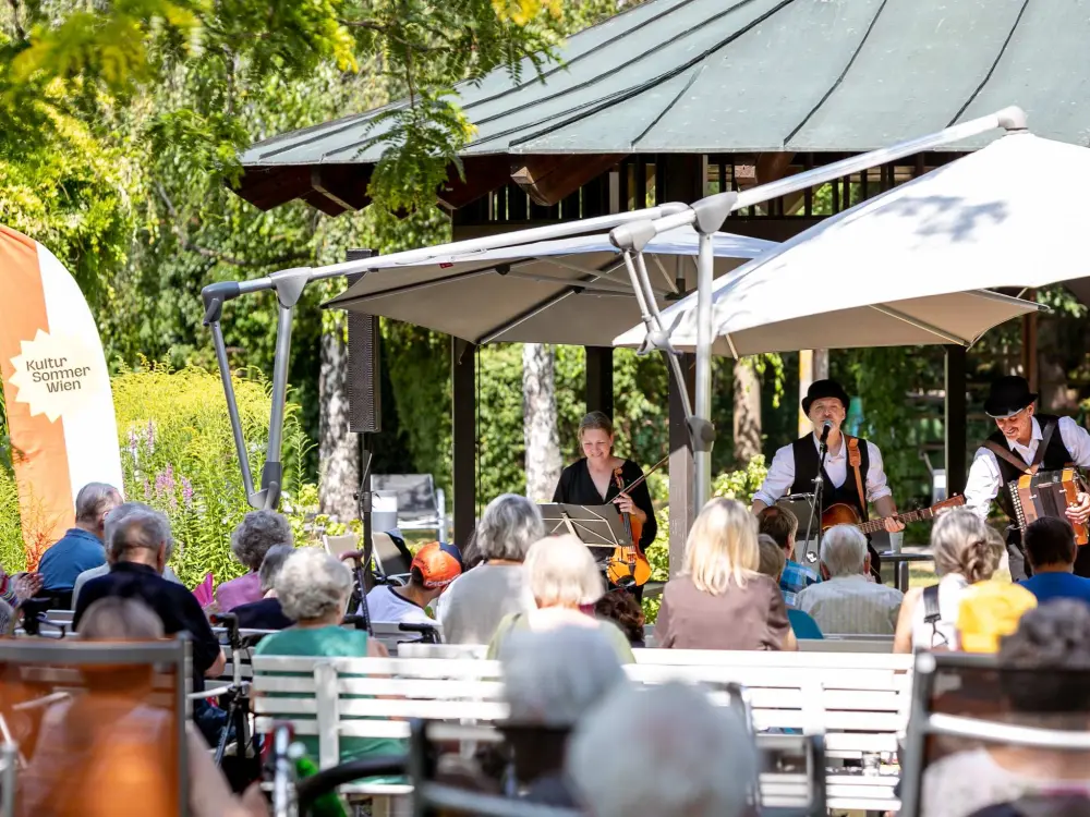 Das Wienerlied-Duo Lipp & Lenz bei ihrem Auftritt im Haus zum Leben Mühlengrund. Sie musizieren in einem kleinen Garten, davor sitzen Menschen in Stühlen und hören zu. 