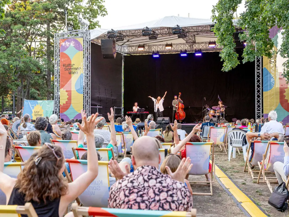 Leléka performt auf der Kultursommer-Bühne. Einige Menschen in den Liegestühlen strecken ihre Hände in die Höhe.