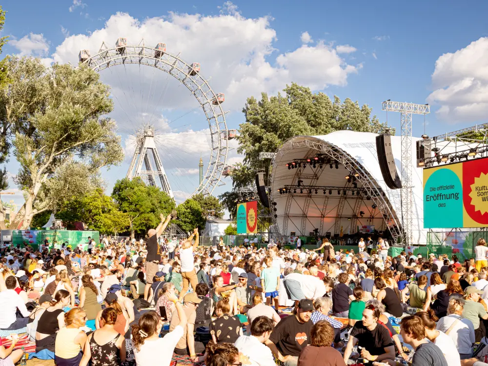 Tausende Menschen sitzen und stehen auf der Kaiserwiese. Rechts im Bild ist eine große Open-Air Bühne, die Wiener Symhoniker spielen eine Konzert. Im Hintergrund ist das Riesenrad.