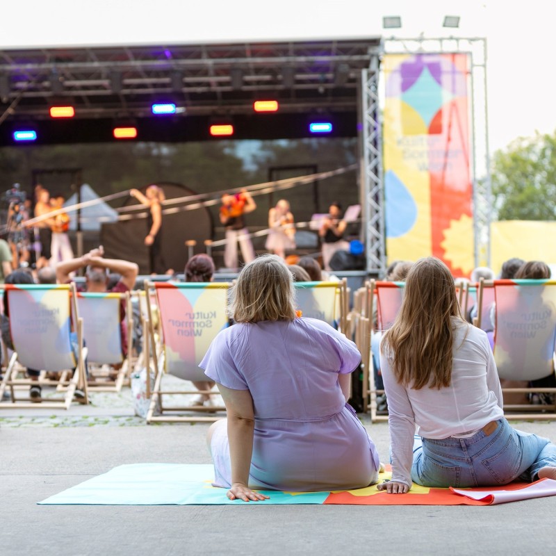 Foto: Judith Stehlik Vor der Bühne am Schrödingerplatz sitzt Publikum in den Liegestühlen. Zwei Frauen sitzen auf einer Decke am Boden und schauen auf die Bühne. Auf der Bühne sieht man das Freestyle Orchestra, sie machen Musik und gleichzeitig Akrobatik.