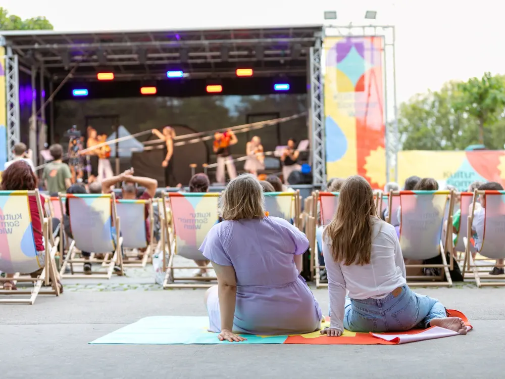 Vor der Bühne am Schrödingerplatz sitzt Publikum in den Liegestühlen. Zwei Frauen sitzen auf einer Decke am Boden und schauen auf die Bühne. Auf der Bühne sieht man das Freestyle Orchestra, sie machen Musik und gleichzeitig Akrobatik.