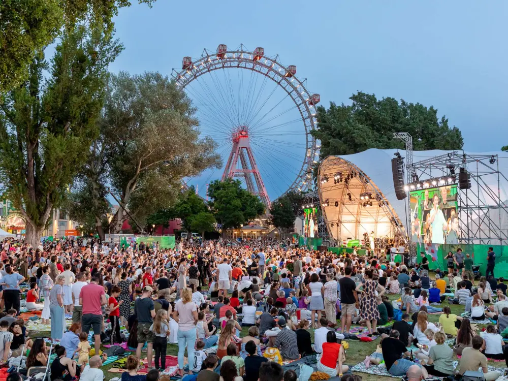 Blick auf die Kaiserwiese. Im Hintergrudn sieht man das Riesenrad. Im Vordergrund stehen und stitzen hunderte Menschen und blicken auf die Bühne mit den Wiener Symphonikern