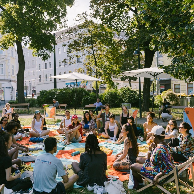 Künstler:innen sitzen im Reithofferpark auf einer Picknickdecke und diskutieren. 