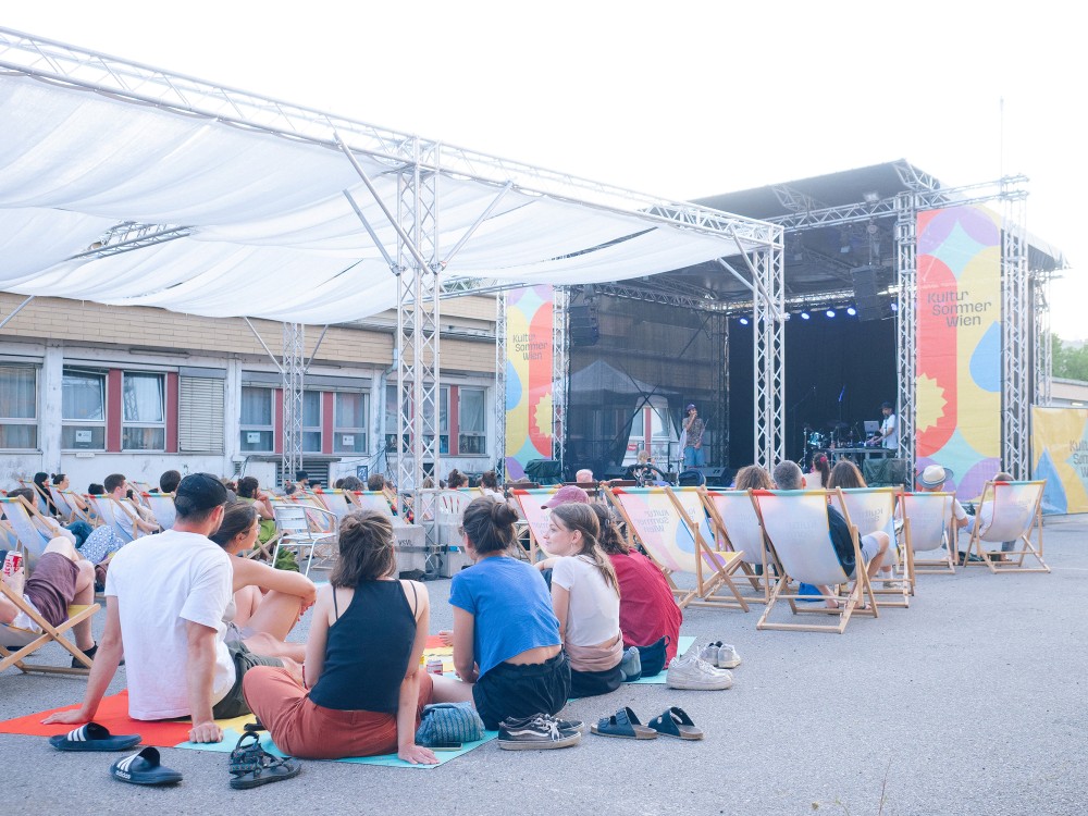 Menschen sitzen in Liegestühlen und auf Picknickdecken vor der Kultursommer-Bühne beim Nordwestbahnhof. Auf der Bühne treten Gewinner:innen des WIENXTRA-Sounbase Wettbewerbs auf. 