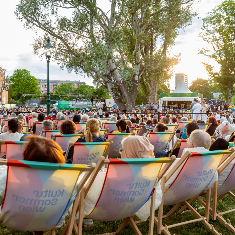Foto: Judith Stehlik Besucher:innen des Pratr-Picknicks der Wiener Symphoniker sitzen in Kultursomner-Liegestühlen. Die Stühle sind Richtung Bühne ausgerüchtet.