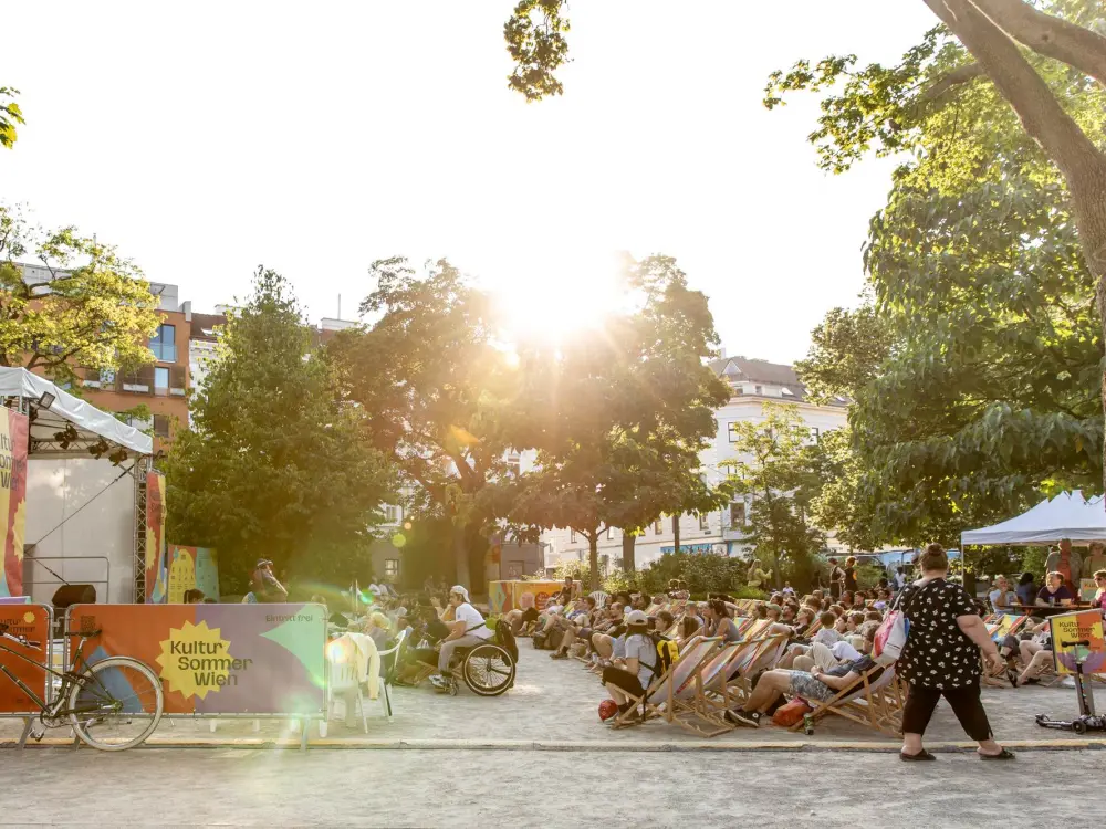 Reithofferpark: Das Publikum und die Kultursommer-Bühne im Reithofferpark von der Seite. Die Sonne strahlt kräftig hinter einem Baum hervor.