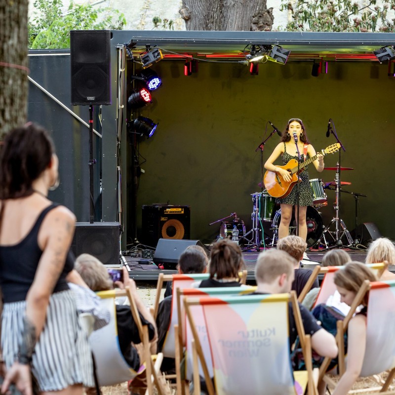 Foto: Judith Stehlik Musikerin Lila mit Gitarre auf der Bühne im Hyblerpark. Vor der Bühne sitzt Pulikum in Liegestühlen.