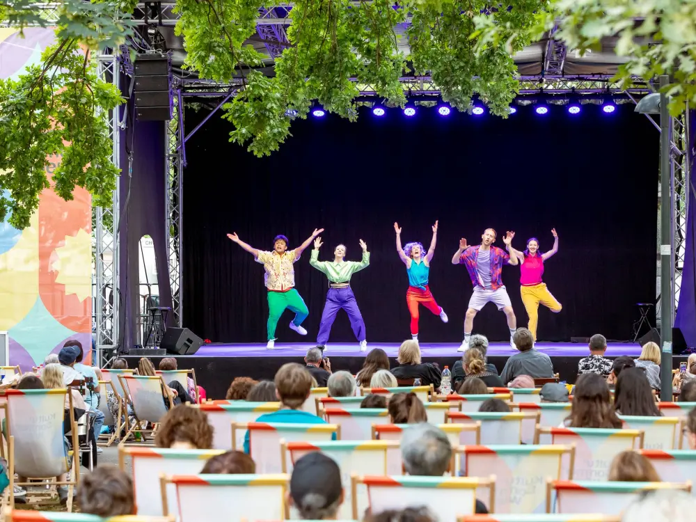 Fünf bunt gekleidete Tänzerinnen und Tänzer springen auf der Bühne in die Luft. Im Vordergrund sitzt das Publikum in Liegestühlen.