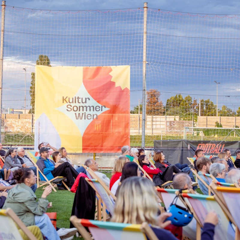 Foto: Judith Stehlik Viele Menschen sitzen in den LIegestühlen des Kultursommer Wein beim Fußballverein 1210. Neben den Liegestühlen ist ein Netz aufgespannt, darauf hängt eine Plane mit dem Kultursommer-Logo. 