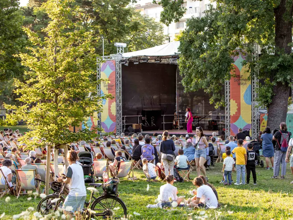 Antonia Stabinger steht auf der Kultursommer-Bühne im Stadtpark Atzgersdorf. Menschen stehen oder sitzen vor der Bühne. Der Park ist gut gefüllt. 