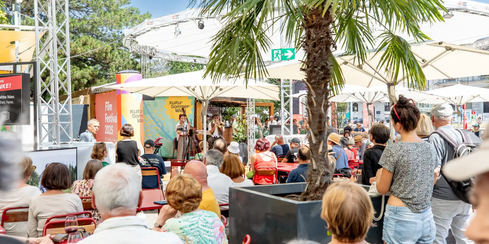 : Menschen sitzen an Tischen vor der Bühne am Rathausplatz.