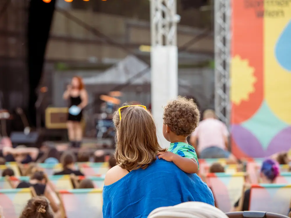 Eine Frau hält ein Kind am Arm. Sie sehen sich Programm auf der Kultursommer Bühne an. 