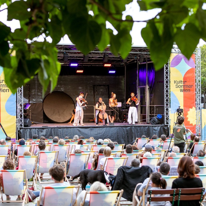 Foto: Judith Stehlik Vor der Bühne am Schrödingerplatz sitzt Publikum in den Liegestühlen. Auf der Bühne sieht man das Freestyle Orchestra, sie machen Musik und gleichzeitig Akrobatik.