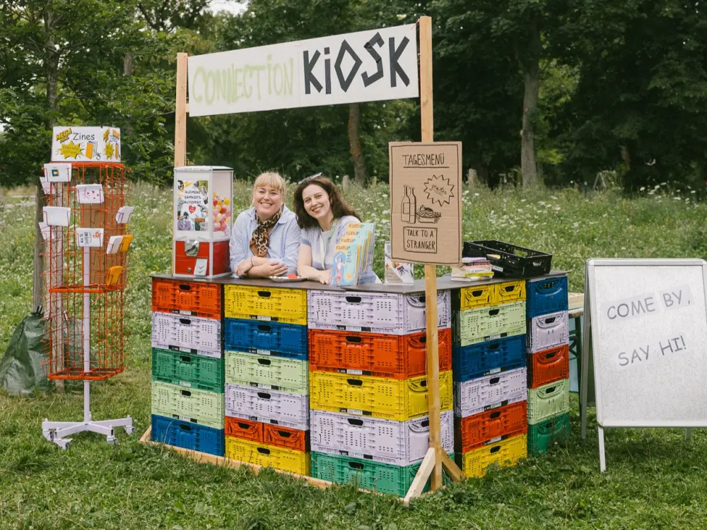 Das Team von Kultursommer Plus steht hinter der Theke des Connection Kiosk-Stand und lächelt in die Kamera. 