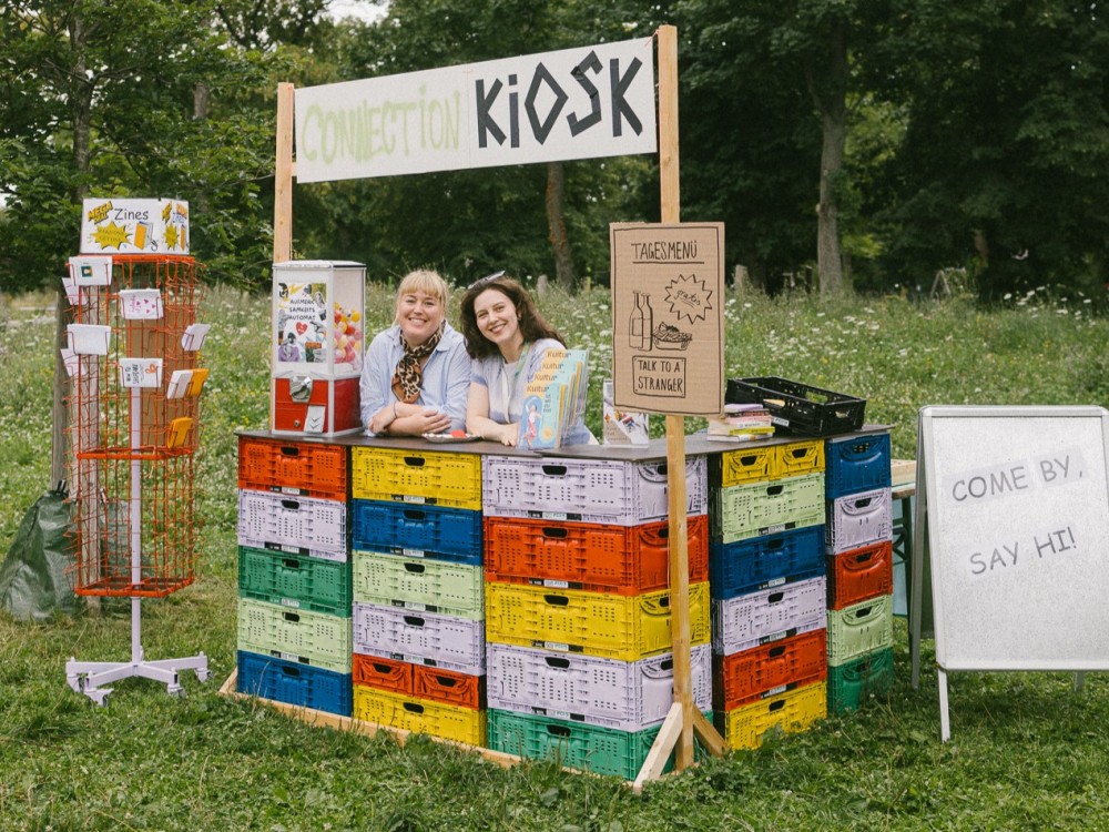 Das Team von Kultursommer Plus steht hinter der Theke des Connection Kiosk-Stand und lächelt in die Kamera. 