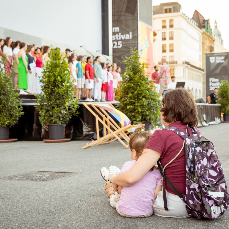 Foto: Judith Stehlik Eine Person sitzt mit einem Kind vor der Bühne am Rathausplatz und hört dem Chor Klangwelle Wien zu.