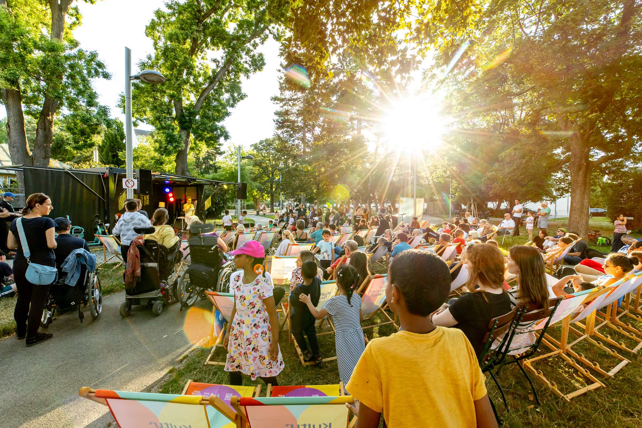 Künstlerin Meo auf der Bühne im Hyblerpark. Man sieht das Publikum von hinten. Kinder und 2 Personen im Rollstuhl sind im Bildvordergrund.