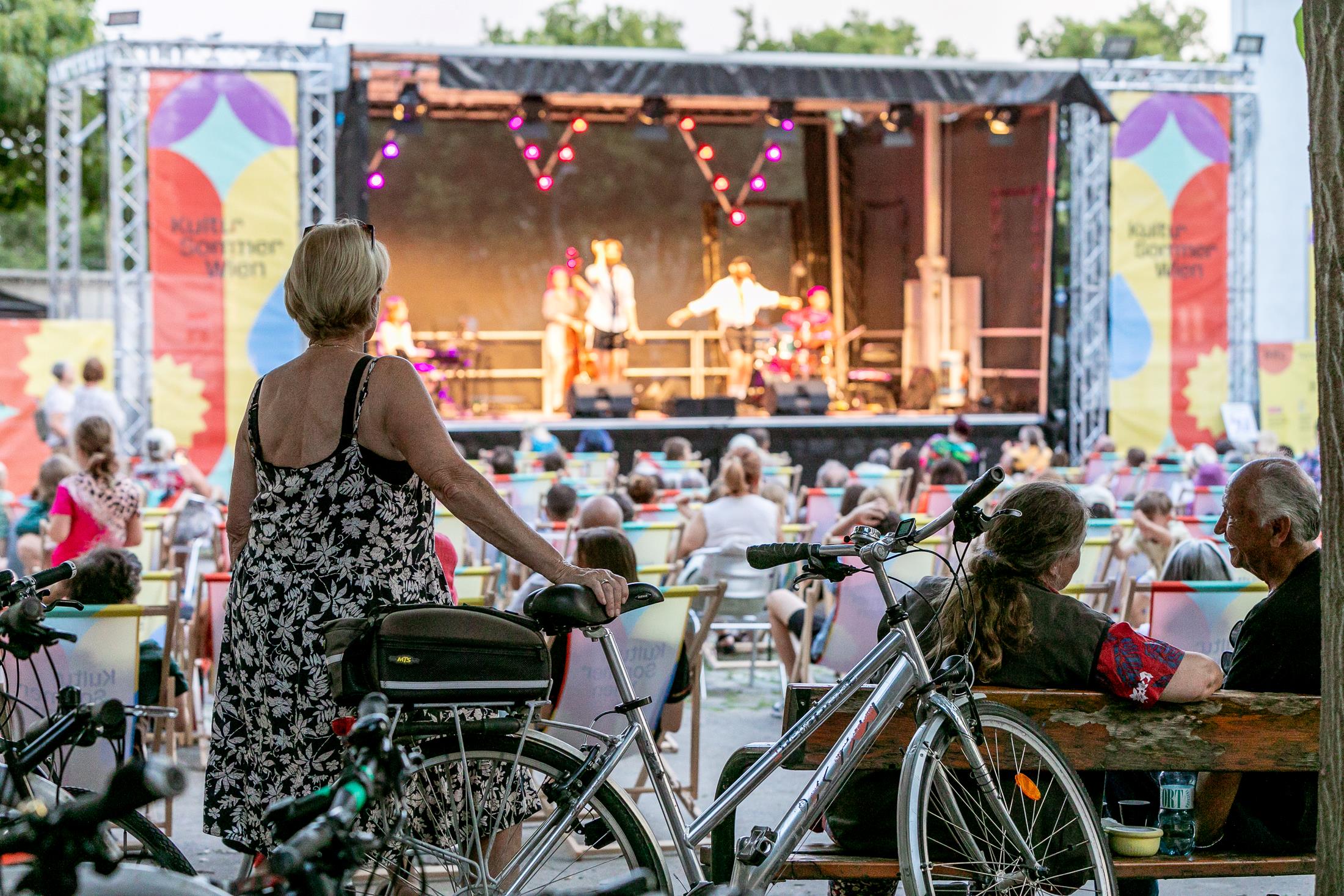 Blick auf die Bühne am Schrödingerplatz: Eine Dame steht mit Fahrad im Vordergrund. Das Publikum sitzt dahinter auf Liegestühlen. Auf der Bühne stehen die Musiker Tom Neuwirth und Martin Zerza mit Band.