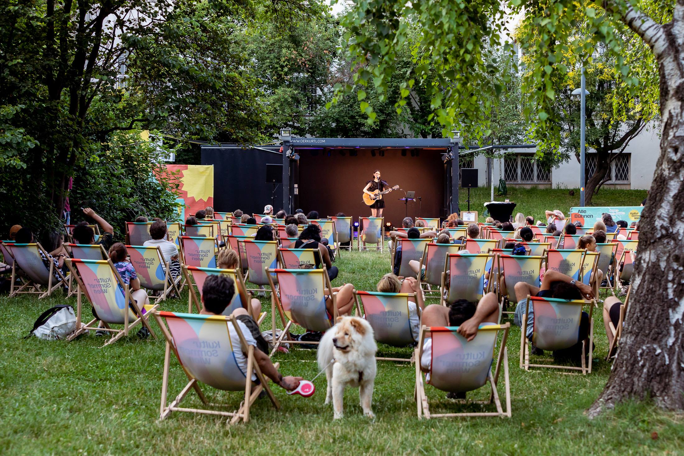 Besucherinnen im Wärhingerpark sitzen in Liegestüheln oder auf Picknickdecken in der Wiese und blicken auf die Bühne. Die Musikerin Mira Taylor steht mit Gitarre in der Hand auf der Bühne.