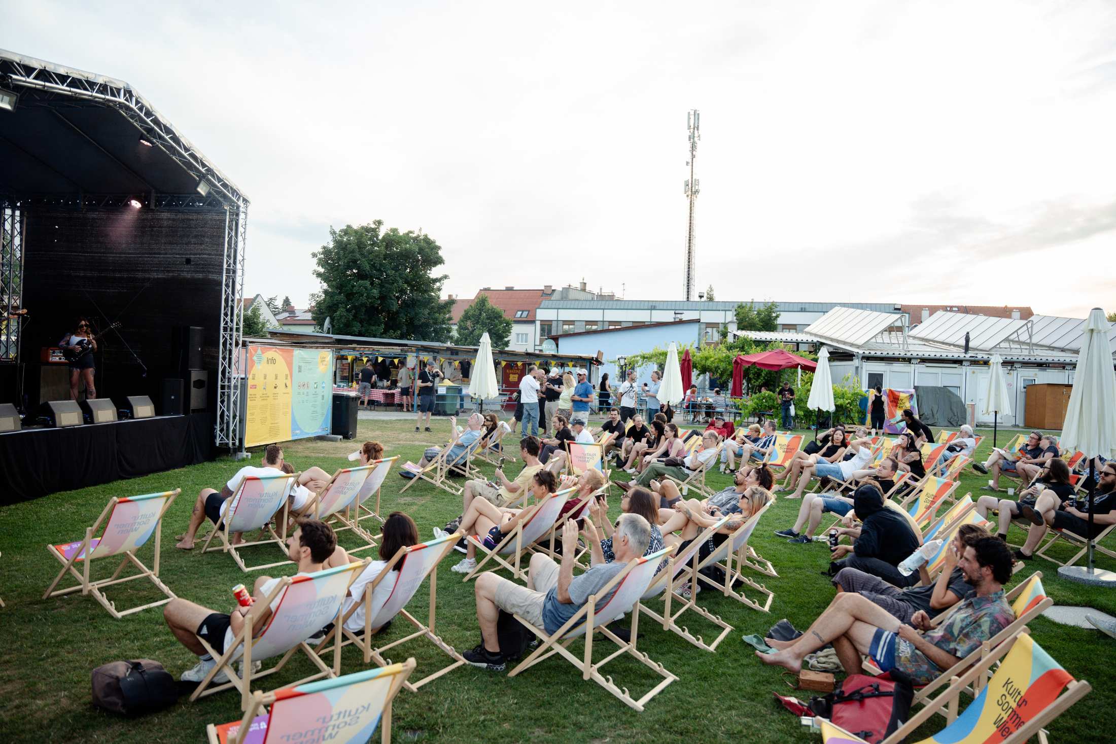 Vor der Kultursommer-Bühne beim Fußballverein 1210 sitzen Menschen in Liegestühlen. 